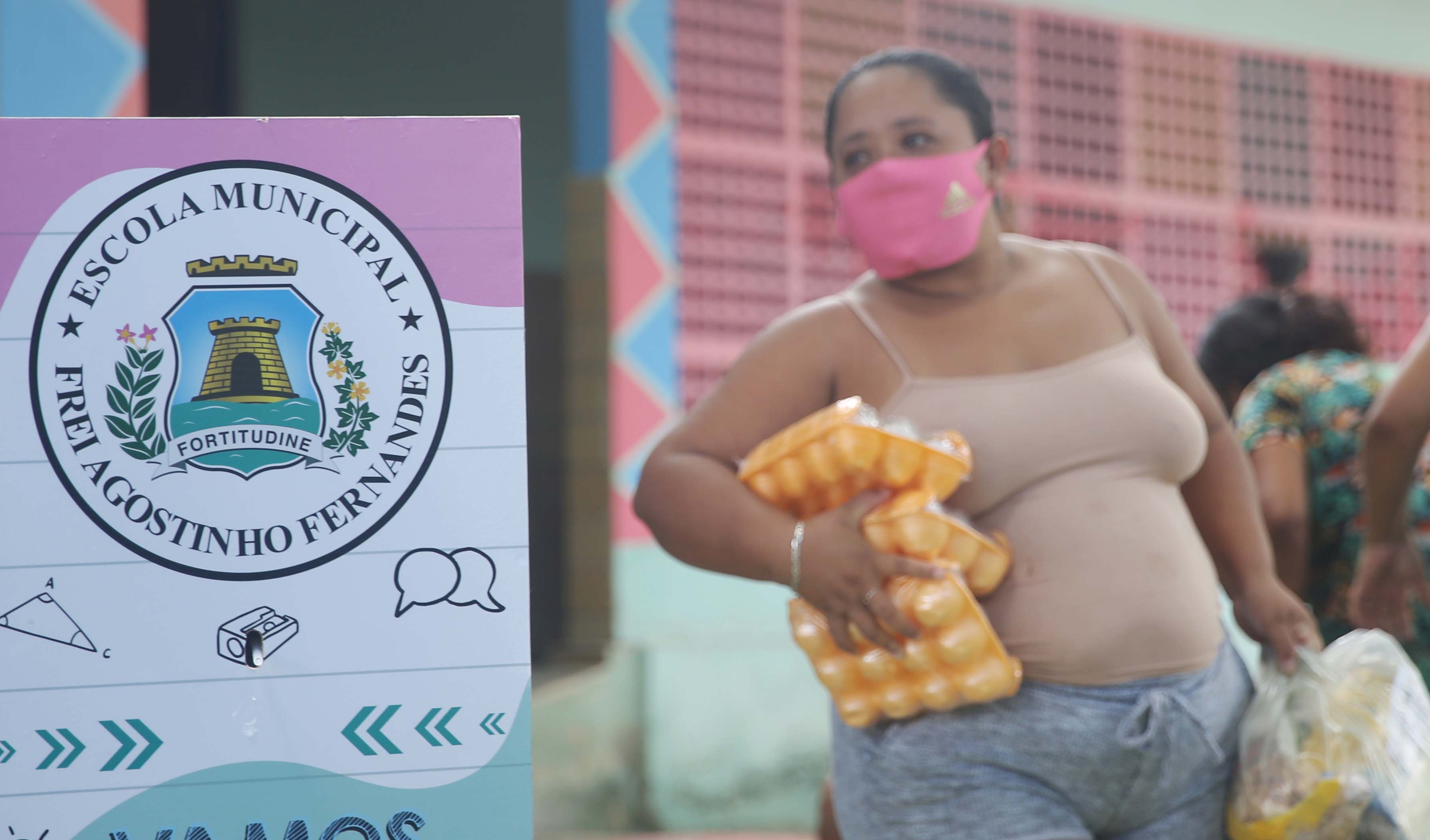 uma mulher de máscara segura um kit de alimentação ao lado de uma placa que orienta as pessoas a lavar as mãos